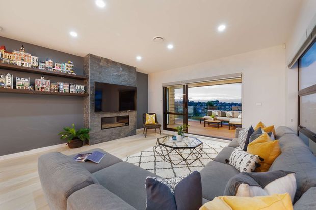 Contemporary living room with a large grey sectional sofa, geometric coffee table, patterned rug, yellow accent cushions, built-in shelving displaying miniature house models, and a stone-clad fireplace feature wall. Full-height sliding glass doors open to a balcony or terrace with a distant urban view.