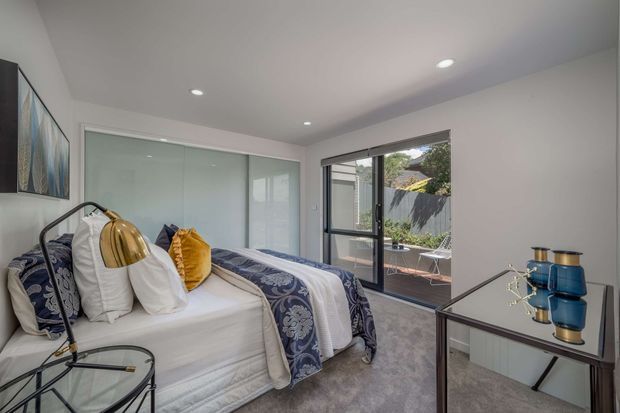 Bright, contemporary bedroom with a neatly made bed, layered white and patterned blue bedding, bedside lamp, framed artwork, mirrored sliding wardrobe doors, and a dark-framed glazed door opening to an outdoor terrace. The room has recessed ceiling lighting, pale walls, and a soft grey carpet.