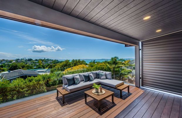 Covered outdoor terrace with timber decking, a built-in ceiling with recessed lights, and a large louvred or roller-style privacy screen. A sectional outdoor sofa and coffee tables face a landscaped suburban view with the ocean visible in the distance.
