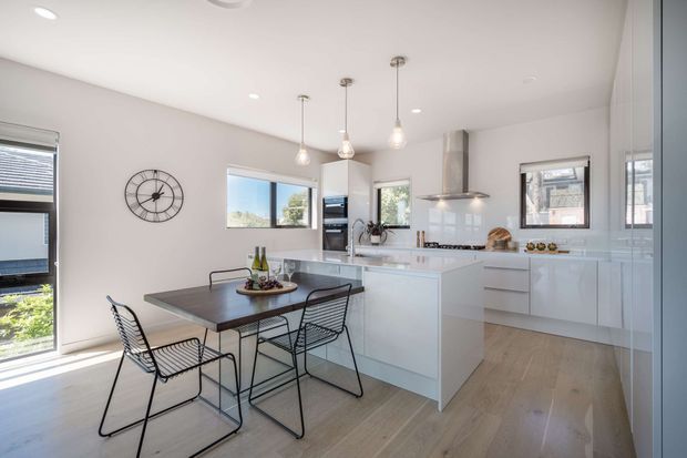 Bright contemporary open-plan kitchen and dining area with white flat-panel cabinetry, a central island, stainless-steel rangehood, light timber flooring, and a dark rectangular dining table with wire-frame chairs. Pendant lights, large windows, and skylights provide abundant natural light.