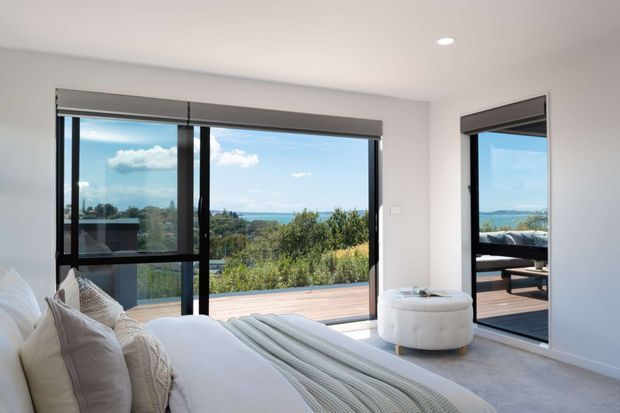Bright contemporary bedroom with a large upholstered bed, soft neutral bedding, a round ottoman, light carpet, and expansive black-framed glazing. The windows open to a timber deck and leafy coastal or waterfront outlook, with a reflected outdoor seating area visible in the right-hand glazing.