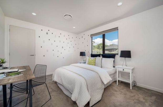 Bright contemporary bedroom with a white upholstered bed, layered white bedding, black bedside lamps, a white bedside table, patterned feature wall decals, grey carpet, and a compact desk with wire chair. A large window provides views of neighbouring rooftops and greenery.