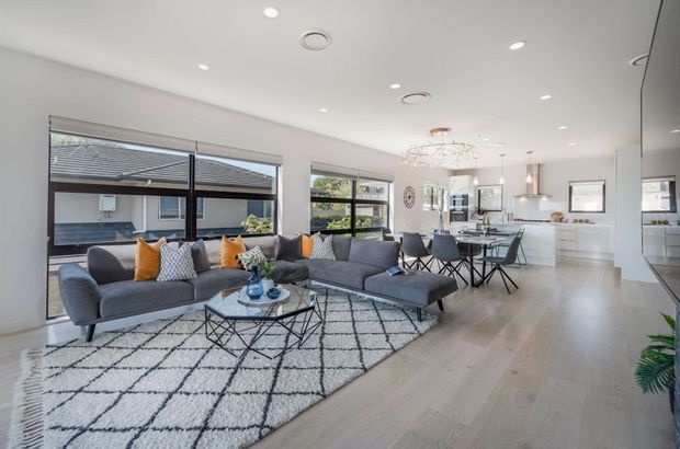 Bright open-plan living and dining area with a contemporary kitchen in the background. The room features a large charcoal sectional sofa, patterned area rug, glass-topped coffee table, dining table with dark chairs, light timber flooring, white walls and ceiling, expansive black-framed glazing, and modern pendant and recessed lighting.