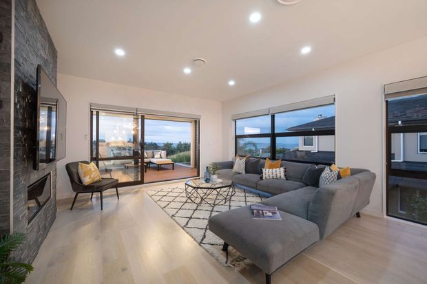 Bright contemporary living room with pale timber flooring, white walls and recessed ceiling lights. A large grey sectional sofa faces a wall-mounted TV and dark stone-clad fireplace, with patterned cushions, a geometric rug and a black wire coffee table. Floor-to-ceiling sliding glazing opens toward an outdoor balcony or terrace, while large windows provide views of neighbouring homes.