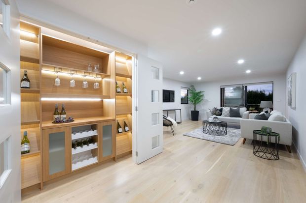 Bright contemporary open-plan living room featuring a built-in timber bar and display cabinet with glass-fronted storage, integrated lighting, pale timber flooring, and a white sectional seating area. Large windows, recessed ceiling lights, and neutral furnishings create an airy, refined entertaining space.