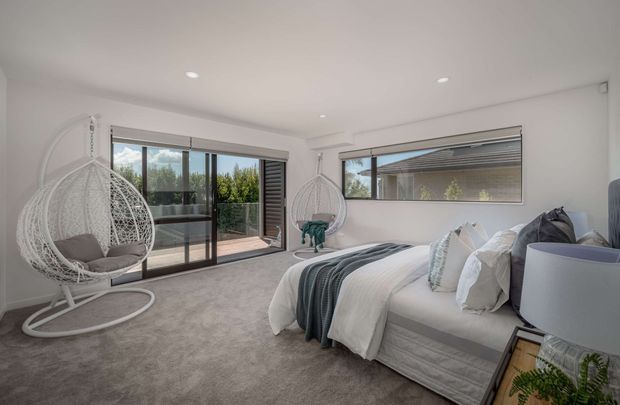 Bright contemporary bedroom with a large upholstered bed, layered white and grey bedding, carpeted flooring, floor-to-ceiling sliding glass doors opening to a balcony or terrace, and a long horizontal window. Two woven hanging egg chairs add a relaxed decorative element, while recessed ceiling lights and neutral finishes create an airy, comfortable interior.