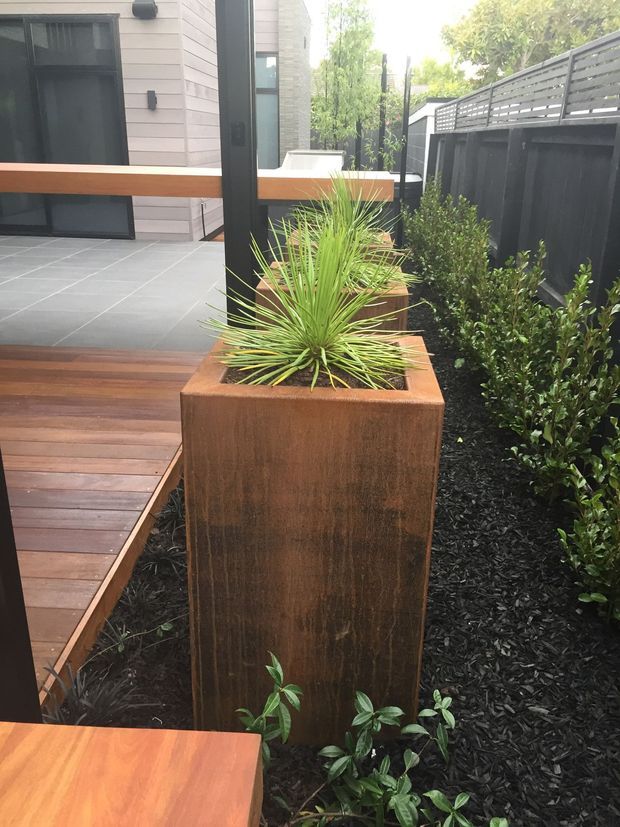 A landscaped courtyard or patio edge beside a modern home, featuring a tall rusted metal planter with spiky ornamental foliage, dark mulch, clipped shrubs, warm timber decking, and large grey floor tiles. A dark horizontal-slat privacy fence and contemporary glazing frame the garden space.