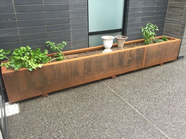 An outdoor architectural planter box made from weathered rust-toned metal, positioned against a dark charcoal tiled wall. The long rectangular planter contains green plants and small decorative pots, with a textured grey paved ground in front.