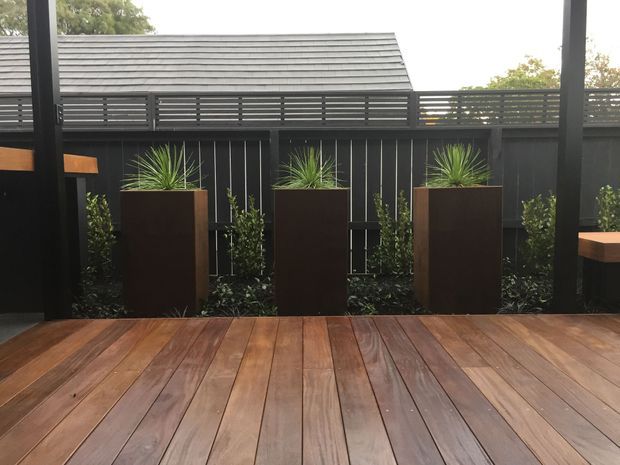 A covered outdoor deck with warm timber decking, dark painted structural posts and slatted privacy fencing. Three tall rectangular planters with spiky green plants line the edge, with a grey tiled pitched roof visible beyond the fence.