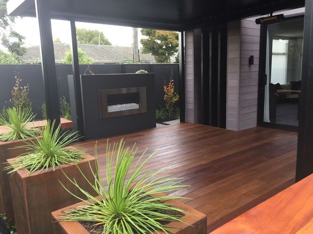 A covered outdoor deck or terrace with rich timber decking, integrated raised planter boxes, and a dark built-in outdoor fireplace or fire feature. The space is enclosed by dark privacy screening and framed glazing, with a doorway opening into the home. Contemporary black and timber finishes create a warm, sheltered outdoor entertaining area.