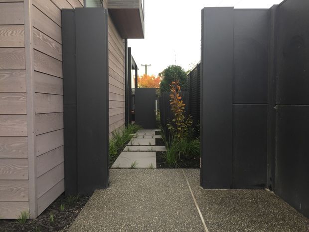 A narrow landscaped side passage between a timber-clad building and a dark metal boundary fence. The path combines exposed aggregate paving with square concrete stepping slabs, black garden edging, and slender ornamental planting. The view leads toward a small rear garden or courtyard area.