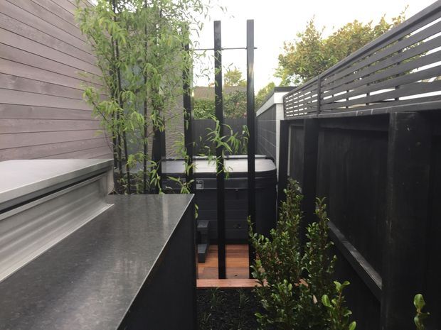 A narrow landscaped outdoor courtyard or side passage with dark horizontal timber screening, black metal posts, raised planter beds, and a compact built-in bench or counter along the left wall. Timber decking and dense vertical planting create a private, contemporary outdoor space.