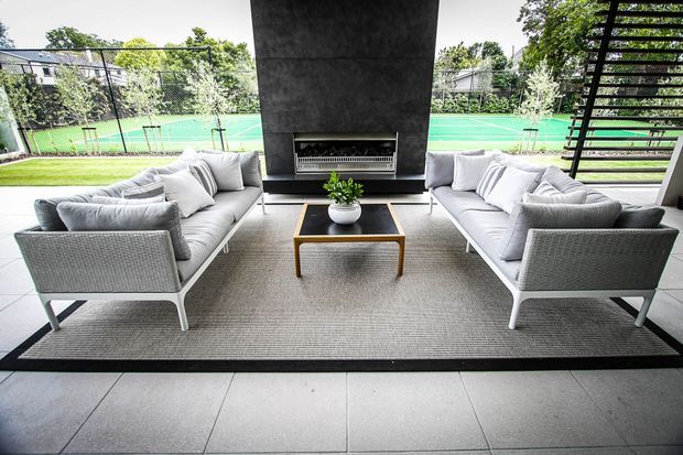A contemporary covered outdoor lounge arranged around a central dark stone or concrete fireplace. Two light-grey sofas face each other across a wood-framed coffee table, with a large neutral rug and tiled flooring. Black slatted screening and glazed openings frame views toward a bright green tennis court and landscaped surroundings.