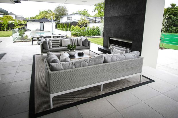 A covered outdoor lounge and entertaining area with two large grey sofas arranged around a low coffee table and a dark built-in fireplace feature wall. Large pale-grey tiled flooring extends toward a landscaped garden, pool area, and green tennis court beyond. The space has a clean, open contemporary design with strong indoor-outdoor connection.