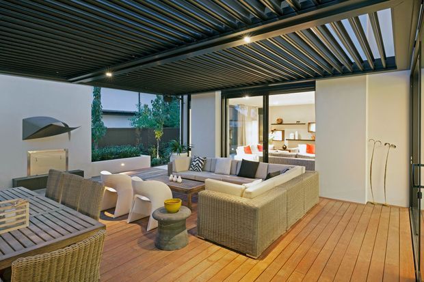 A covered outdoor living and entertaining area with timber decking, woven outdoor lounge furniture, a dining table, and a large louvred pergola roof. The space opens to a landscaped courtyard and connects visually to a bright interior room through expansive glazing. Neutral woven furnishings and warm timber create a relaxed, contemporary atmosphere.