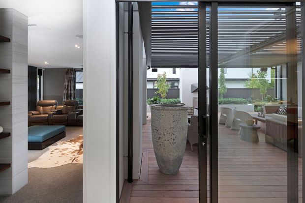 View from an interior living area through full-height dark-framed glazed doors to a covered timber deck and landscaped courtyard. The space features a large sculptural planter, slatted pergola overhead, outdoor seating, and a glimpse of a contemporary lounge with built-in shelving and leather seating.