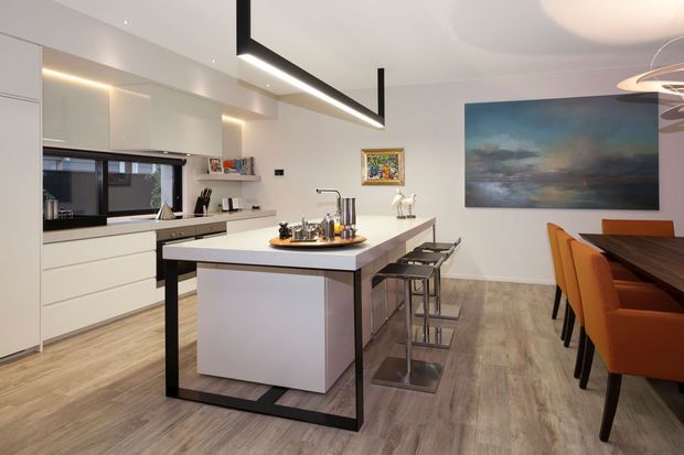 A bright contemporary open-plan kitchen and dining area with white handleless cabinetry, a large central island, integrated appliances, and black-framed linear pendant lighting. The island has a white benchtop with dark metal framing and stools along one side. A dark timber dining table with orange upholstered chairs sits beside the kitchen, while a large abstract artwork provides a blue-green accent on the white wall. Light wood-look flooring runs throughout.