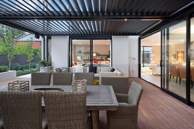 A covered outdoor entertaining terrace with a large wicker dining table and chairs, lounge seating, timber decking, and a black louvred pergola overhead. The terrace opens directly to a bright contemporary interior through expansive sliding glass doors, with landscaped planting and a privacy fence visible at the edge.