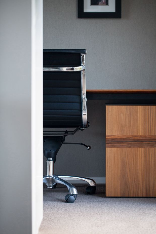A compact, refined home office viewed through a doorway, featuring a black leather office chair with chrome frame and casters, a dark built-in cabinet with warm timber drawer fronts, grey textured walls, and grey carpet. The space has a restrained mid-century-inspired palette and clean built-in joinery.