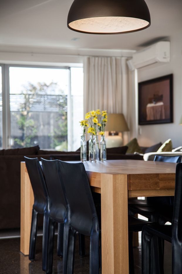 A warm contemporary open-plan dining and living area with a substantial light timber dining table surrounded by black leather chairs. A dark pendant light hangs overhead, while a sofa, floor lamp, artwork, curtains, and large glazed doors create a comfortable residential setting. Natural daylight enters from the rear glazing, with an air-conditioning unit mounted high on the wall.