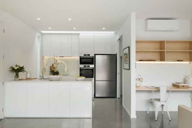 Bright open-plan kitchen and compact home office in a clean contemporary interior. The kitchen features glossy white flat-panel cabinetry, a central island with sink, integrated appliances, stainless-steel refrigerator, pale stone or composite benchtops, and a softly illuminated backsplash. Beside it, a built-in timber-toned shelving and desk area provides a small workspace with a white metal chair. The space has polished grey flooring, recessed ceiling lighting, and a wall-mounted split-system air conditioner.