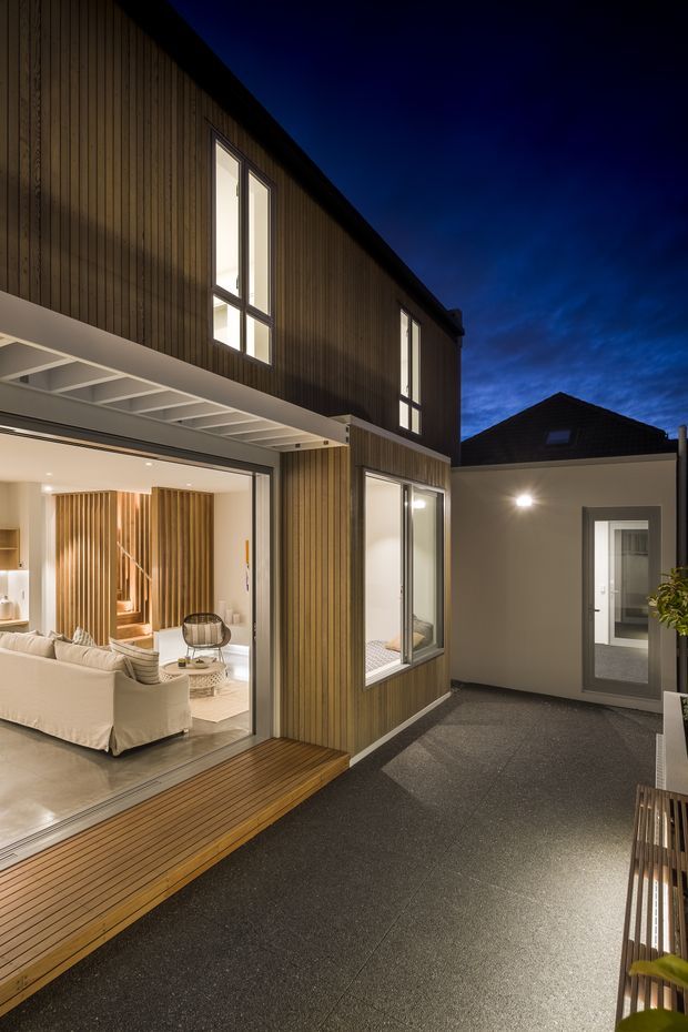 Twilight exterior view of a contemporary multi-level home surrounding a paved courtyard or terrace. The facade combines vertical timber cladding, large glazed openings, a projecting covered outdoor area, and dark roof forms. Warm interior lighting reveals an open-plan living room with pale sofas and timber feature screening through the wide sliding glass doors.