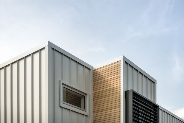 Exterior architectural view of a contemporary angular building with light grey vertically ribbed metal cladding, a contrasting warm timber-clad recessed wall, a small horizontal window, and a dark horizontal louvred screen. The composition emphasizes intersecting rooflines and facade planes against a pale sky.