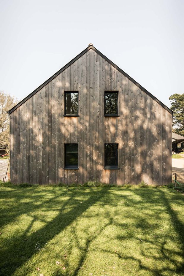 Front exterior view of a simple two-storey gable-ended building clad in vertically arranged weathered timber boards. Four black-framed rectangular windows are symmetrically arranged on the facade, with a dark roof edge and lawn in the foreground. Mature trees cast dappled shadows across the timber facade and grass.