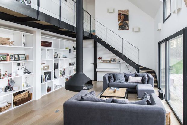 Bright, spacious contemporary living room with a double-height void and mezzanine walkway. The room features white built-in shelving, a black steel staircase with slim balustrade, a suspended black freestanding fireplace, two dark grey sofas, and a timber coffee table. Large windows and glazing bring in abundant natural light, while framed artwork and display objects add visual interest.