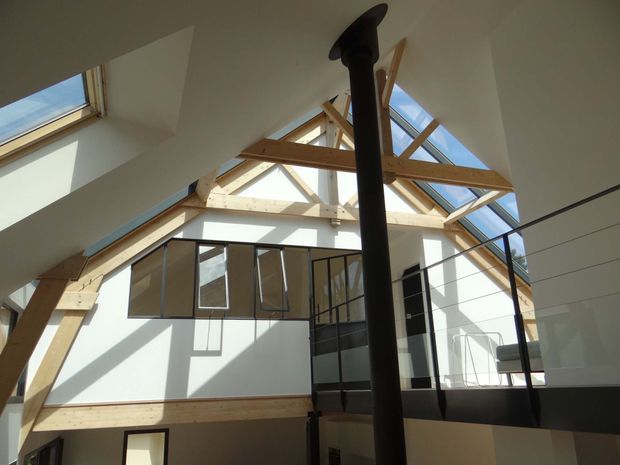 Interior view of a bright, multi-level open-plan space with a high pitched roof and exposed timber post-and-beam structure. Large skylights and clerestory glazing bring in strong daylight, while black-framed glass balustrades and a central black flue add contrast. A mezzanine or upper landing overlooks the lower level.