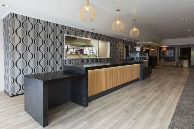 A contemporary hospitality or commercial interior featuring a long reception or service counter with a black stone-look benchtop, black cabinetry, and a vertical timber slat front. The space has patterned geometric wallpaper, light timber-look flooring, pendant lights with woven or ribbed shades, recessed ceiling lighting, and a bar or service area visible behind the counter.