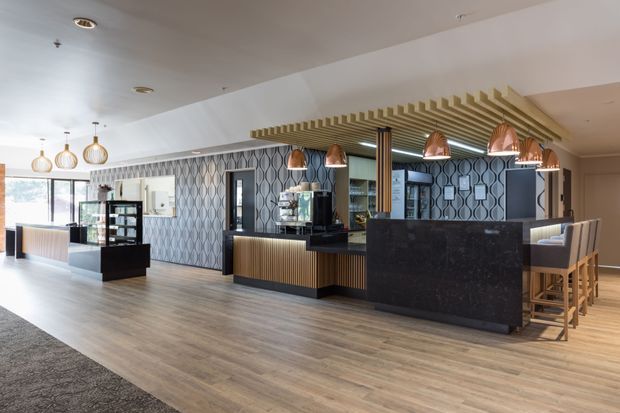 A spacious contemporary hospitality-style interior featuring a bar or reception counter with dark stone surfaces, warm timber slatted ceiling detailing, patterned feature wallpaper, pendant lighting, display shelving, and high stools. Large windows brighten the open-plan space, with timber-look flooring extending across the room.