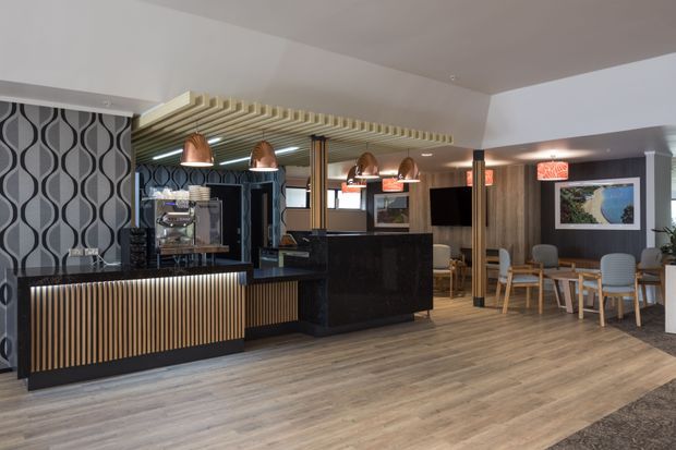 A contemporary hospitality interior featuring a café or bar counter with ribbed timber panelling, black stone surfaces, patterned feature wallpaper, exposed timber ceiling battens, and multiple copper pendant lights. The space includes a dining area with timber tables and upholstered chairs, dark timber wall cladding, framed artwork, and wide timber-look flooring.