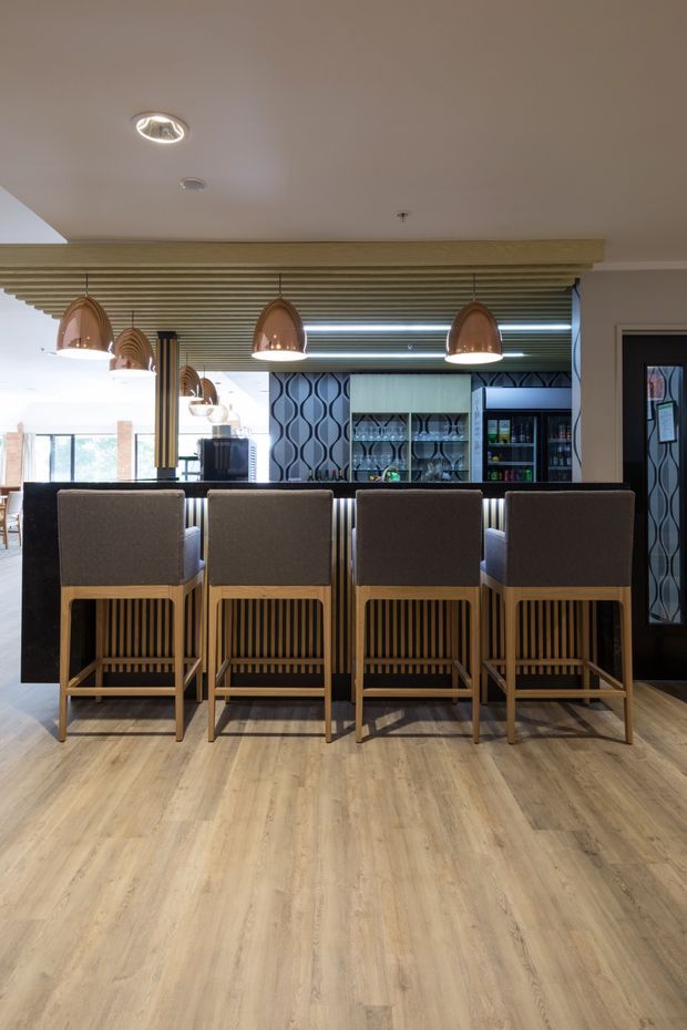 A refined bar or kitchen entertaining area viewed from the seating side, with four upholstered timber bar stools at a dark counter. The space features a slatted timber ceiling detail, warm pendant lights, patterned decorative wall panels, glazed display shelving, dark cabinetry, and light timber-look flooring.