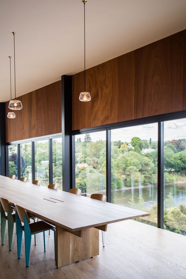 A refined dining room with a long light-wood dining table and matching integrated central support, surrounded by simple upholstered chairs. Floor-to-ceiling black-framed glazing overlooks a leafy landscape and water beyond. The room features rich dark timber wall panelling, pale flooring, a white ceiling, and multiple suspended pendant lights with warm brass details. The composition is warm, modern, and strongly connected to the outdoors.