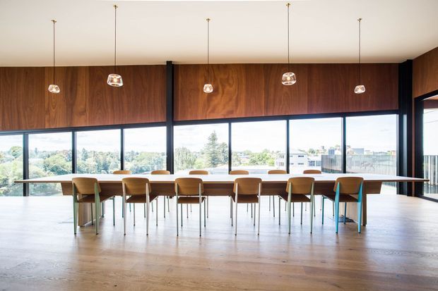 A spacious, bright dining and meeting room with a long timber table surrounded by simple chairs. Floor-to-ceiling windows provide expansive views, while dark timber wall panelling, slim black structural framing, and evenly spaced pendant lights create a refined contemporary interior.