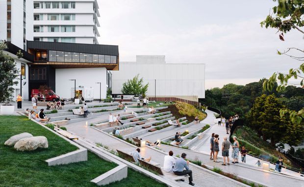 A landscaped multi-level outdoor terrace and amphitheatre beside a contemporary multi-storey building. Broad concrete steps and seating terraces are arranged around planted garden beds, with people using the space. The building combines white rendered volumes, extensive glazing, and a dark metal-clad projecting upper level. Dense mature greenery is visible beyond the site.