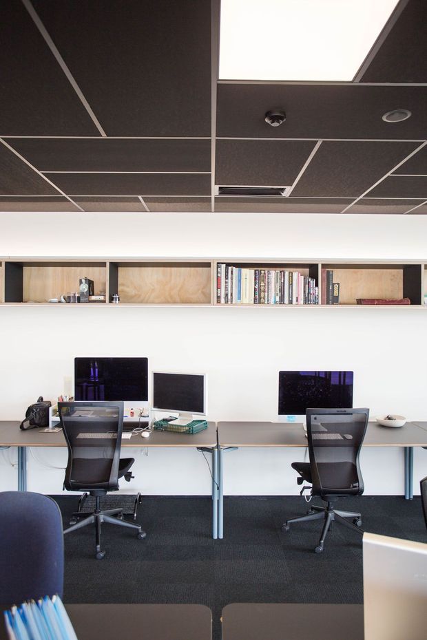 A compact, contemporary shared office workspace with two computer workstations arranged along a long desk. Black ergonomic mesh chairs, monitors, keyboards, and office accessories sit against a white wall. A recessed timber-lined shelving unit displays books and small objects above the desk. The ceiling uses dark acoustic tiles in a regular grid with recessed lighting, ventilation, and a large illuminated panel. Dark carpet and neutral furnishings create a practical, focused atmosphere.