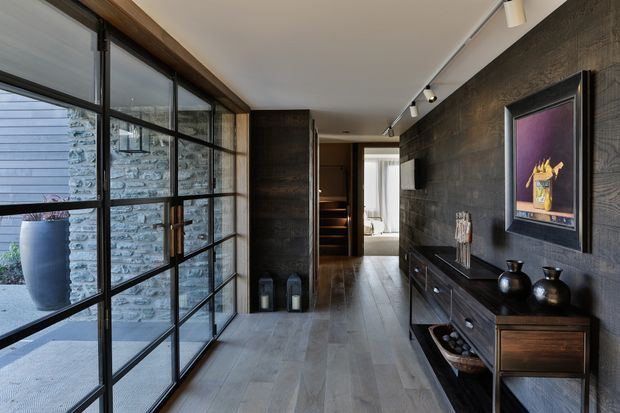 A refined modern entry hall with full-height black-framed glass doors opening to a stone courtyard. The space features dark timber wall panelling, pale timber-look flooring, a slim timber console with drawers, track lighting, and a framed artwork. A corridor leads toward a staircase and brighter interior spaces.
