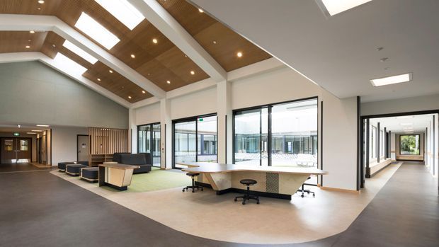 A spacious contemporary commercial interior featuring an open-plan reception or collaborative workspace, lounge seating, shared desks, extensive glazing, and a dramatic timber-lined vaulted ceiling with skylights. Neutral finishes, exposed structural framing, and a long corridor create a bright, refined workplace atmosphere.
