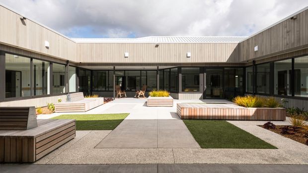 A contemporary single-storey U-shaped building enclosing a landscaped courtyard. The facade combines light vertical timber cladding, dark-framed glazing, and a low pale metal roof. Concrete paths and paving divide rectangular areas of artificial turf and gravel, with integrated timber planter benches containing ornamental grasses.