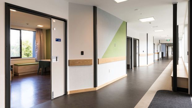 A bright contemporary institutional hallway with dark flooring, white walls, black-framed doorways, timber wall rails, and recessed ceiling lights. The corridor includes multiple rooms and doors, with a glimpse of a furnished bedroom or patient room through the open doorway on the left. A geometric green and grey wall feature adds visual interest.