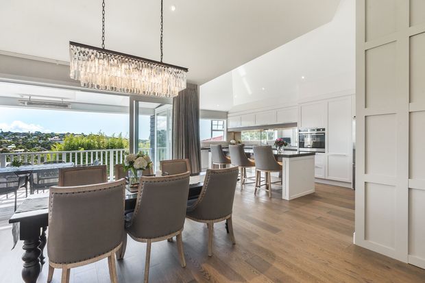 Bright open-plan dining and kitchen interior with a long rectangular crystal pendant above a timber dining table and upholstered dining chairs. White cabinetry, a kitchen island, integrated appliances, warm timber flooring, wall panelling, and expansive glazing create a refined contemporary space. The glazing opens to a balcony or terrace with a leafy suburban outlook.