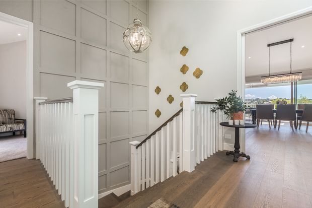 Bright, refined residential hallway and stair landing with a white painted balustrade, square-panel wall moulding, warm timber flooring, and a decorative globe pendant. The stair hall opens toward a formal dining room with a large linear chandelier and expansive glazing.