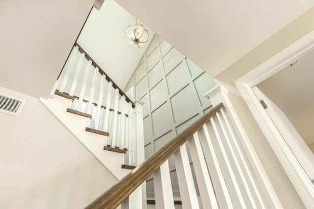 Upward-looking view of a bright residential stairway with white vertical balusters, warm timber handrails and stair treads, ornate white trim, and a tall feature wall with geometric panel moulding. A decorative pendant light hangs above the stairwell, emphasizing the high vertical volume.