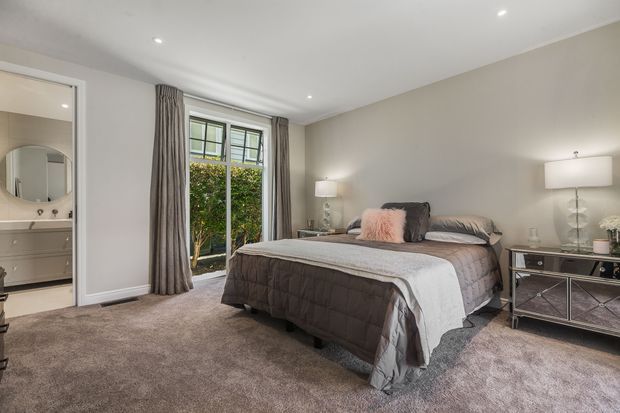 A spacious, softly styled bedroom with a large upholstered bed, layered grey and white bedding, bedside lamps, mirrored bedside cabinetry, full-height curtains, and plush wall-to-wall carpeting. An open doorway reveals a bathroom with a vanity and round mirror. The neutral palette and recessed ceiling lights create a calm, refined atmosphere.