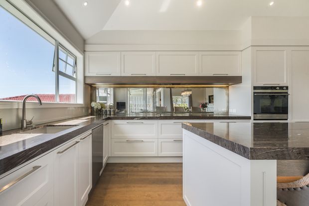 Bright contemporary kitchen with white shaker-style cabinetry, a large central island, dark natural-stone benchtops, mirrored splashback, timber flooring, integrated appliances, and expansive windows. The space has a clean, refined fit-out with warm timber and stone accents.