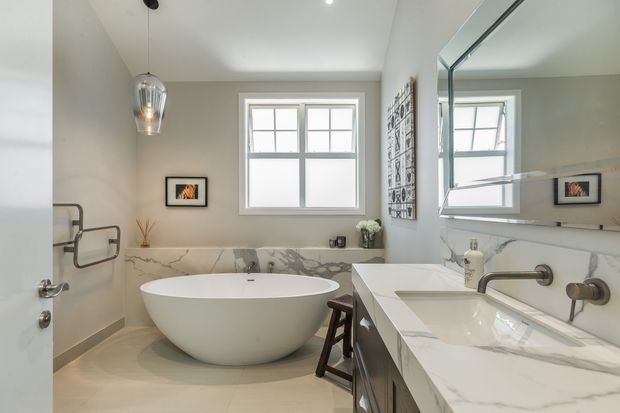 Bright, refined bathroom with a freestanding white oval bathtub, large marble-look wall and vanity surfaces, wall-mounted brushed metal tapware, a double vanity, frameless mirror, and a traditional-style divided-pane window. Neutral finishes and pendant lighting create a clean, contemporary-classic atmosphere.