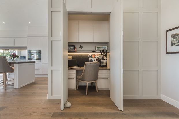 A compact built-in home office nook set within a paneled wall enclosure, featuring a desk, shelving and cabinetry, task lighting, and a upholstered chair. The surrounding interior has white painted wall panelling, warm timber flooring, and an adjacent bright kitchen or dining area visible to the left.