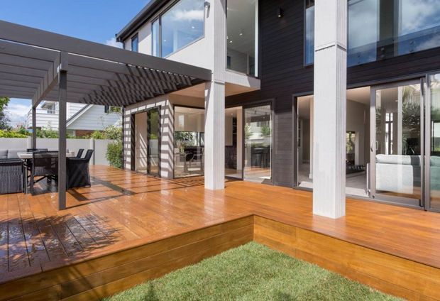 Exterior view of a contemporary multi-level home with a large timber deck, covered outdoor dining area, vertical structural posts, dark horizontal cladding, extensive glazed doors and windows, and an adjoining lawn. Interior living spaces are visible through the glazing.